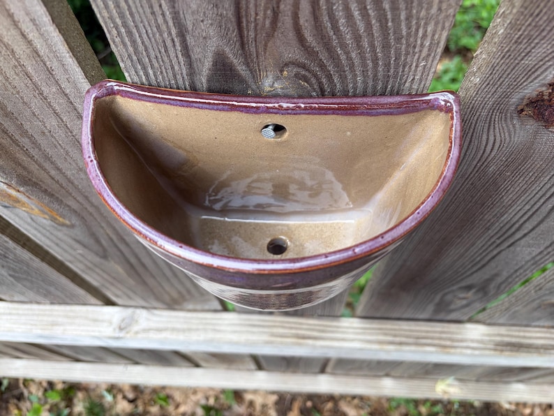 May include: A semi-circular wall-mounted ceramic planter with a glossy tan interior and a dark reddish-brown rim. The piece features a flat back with a pre-drilled hole for mounting and a drainage hole at the bottom of the basin for plant care.