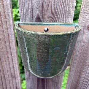 May include: A semi-circular wall-mounted ceramic planter featuring a glossy, deep forest green glaze with subtle variations in tone. The interior reveals a natural, unglazed tan clay surface, and a single circular hole is centered at the top edge.