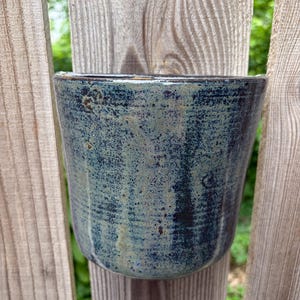 May include: A cylindrical ceramic planter featuring a mottled glaze with deep blue, dark charcoal, and muted sage green tones. The surface has a textured, speckled appearance with subtle vertical striations and a smooth, glossy finish throughout.