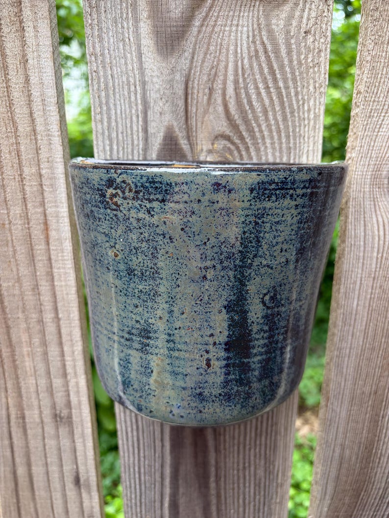 May include: A cylindrical ceramic planter featuring a mottled glaze with deep blue, dark charcoal, and muted sage green tones. The surface has a textured, speckled appearance with subtle vertical striations and a smooth, glossy finish throughout.