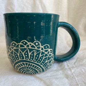 May include: Teal ceramic mug with a white mandala design. The mug has a rounded handle.