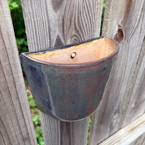 May include: A semi-circular wall-mounted ceramic planter featuring a glossy glaze with blended tones of deep blue, charcoal, and hints of rust red. The interior is a smooth, light tan clay finish with a single pre-drilled hole for mounting.