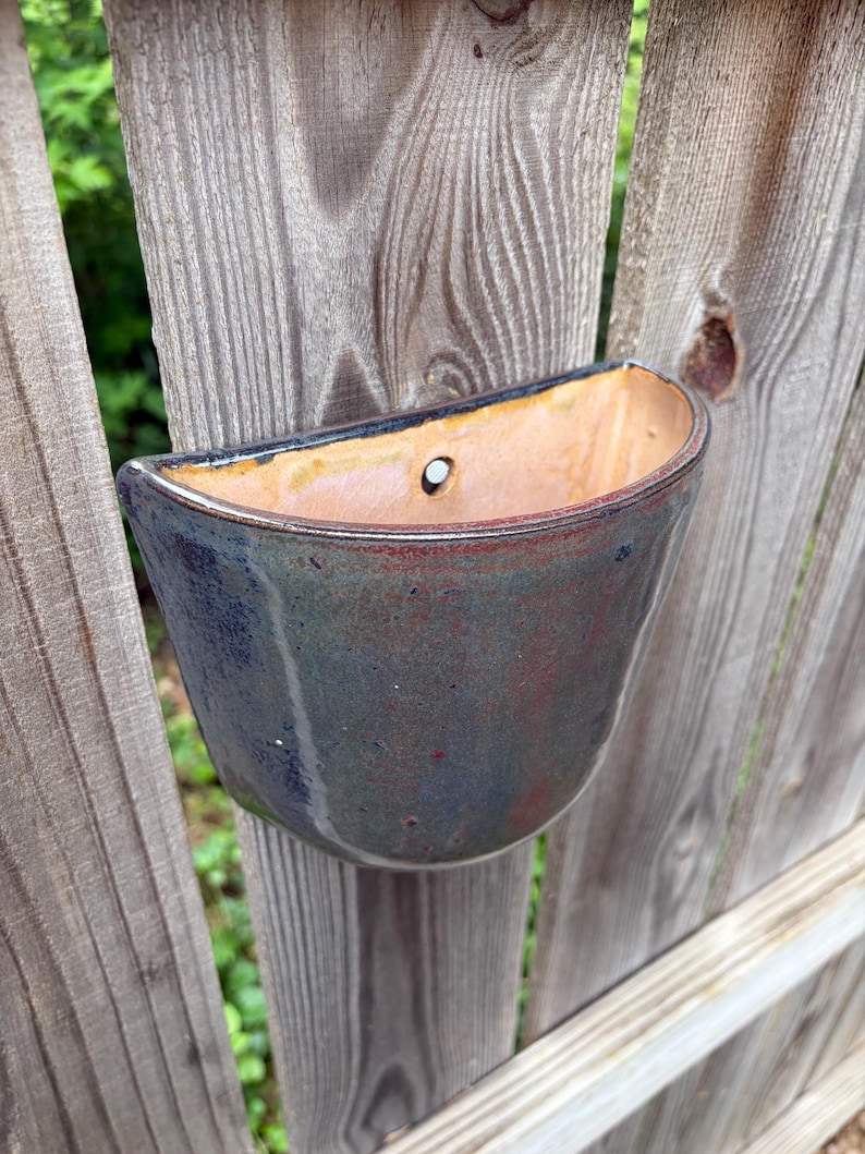 May include: A semi-circular wall-mounted ceramic planter featuring a glossy glaze with blended tones of deep blue, charcoal, and hints of rust red. The interior is a smooth, light tan clay finish with a single pre-drilled hole for mounting.