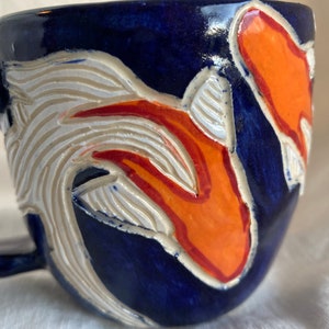 Pu&ograve; includere: Tazza in ceramica con smalto blu scuro. La tazza presenta un design intagliato con accenti bianchi e arancioni. Il manico &egrave; piccolo e curvo. Il design sembra essere un pesce koi.