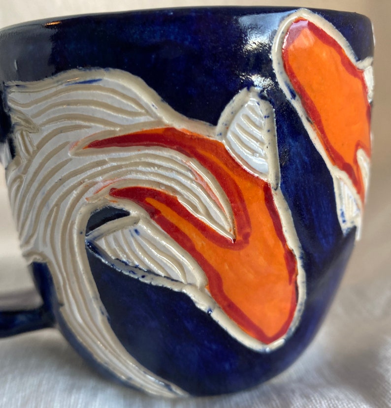 Pu&ograve; includere: Tazza in ceramica con smalto blu scuro. La tazza presenta un design intagliato con accenti bianchi e arancioni. Il manico &egrave; piccolo e curvo. Il design sembra essere un pesce koi.