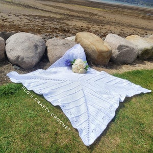 May include: White crochet baby blanket with a scalloped edge, laid out on green grass with a bouquet of white flowers. The blanket has the text "Flo's Crafty Crochet" written on it.