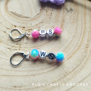 May include: Two sets of colorful beaded stitch markers with silver letter charms spelling "RS" and "WS". The markers are on a wooden surface with a purple crocheted piece and knitting needles.