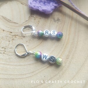May include: Two silver-toned earrings with lever-back closures. Each earring features three beads: two iridescent and one with a letter. One earring has "R S" and the other has "W S". A crochet project and wooden crochet hook are in the background.