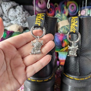 Könnte beinhalten: Ein silberfarbener Metallanhänger in Form eines Hochlandrindes, befestigt an einem runden Ring an der schwarzen Fersenzuglasche eines schwarzen Lederstiefels. Die Zuglasche zeigt den gelben Schriftzug AirWair. Der Anhänger hängt an einem silbernen Metallring.
