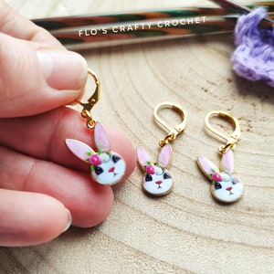 May include: Gold leverback earrings with a white bunny face, pink ears, and a floral crown. The earrings are enamel and have a leverback closure. The text "Flo's Crafty Crochet" is visible in the background.