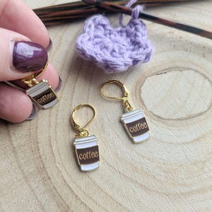 May include: Gold-coloured coffee cup earrings with white and brown enamel detailing. The word "coffee" is printed on the front of the earrings. A small, light purple crocheted heart and crochet hook are in the background. The text "FLO'S CRAFTY CROCHET" is at the bottom.