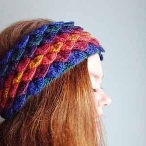 Puede incluir: Una diadema de crochet colorida con un patrón de escamas en tonos de azul, morado, rojo, naranja y amarillo. La diadema la lleva puesta una persona con el pelo largo y castaño.