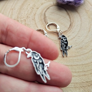 Puede incluir: Pendientes de cuervo plateados con detalles de esmalte negro y blanco. Cada pendiente presenta un cuervo posado en una rama, con un diseño de media luna. El texto "FLO'S CRAFTY CROCHET" es visible.