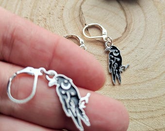 May include: Silver-tone raven earrings with black and white enamel detailing. Each earring features a raven perched on a branch, with a crescent moon design. The earrings are displayed against a wooden background, with the text "FLO'S CRAFTY CROCHET".