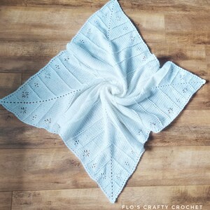 May include: A white crocheted baby blanket with a delicate floral pattern. The blanket is folded and displayed on a wooden surface.