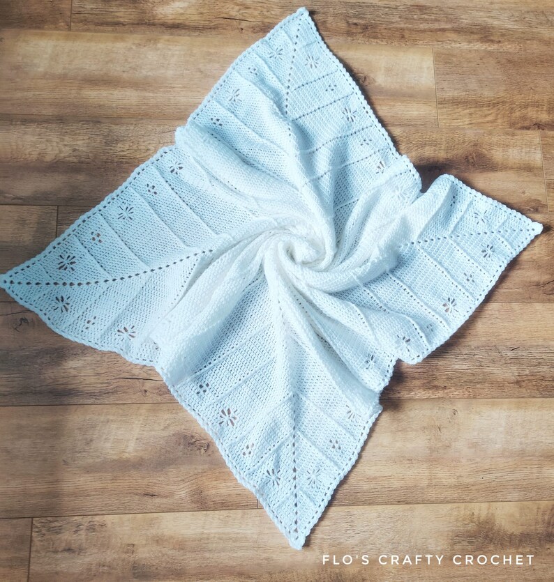 May include: A white crocheted baby blanket with a delicate floral pattern. The blanket is folded and displayed on a wooden surface.