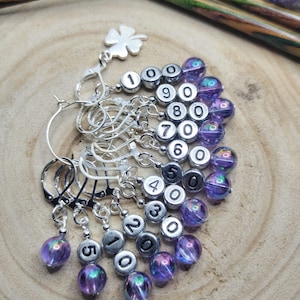 May include: A set of ten silver stitch markers with purple iridescent beads. Each marker has a silver ring and a bead with a black number from 1 to 10. The markers are arranged in a fan shape on a wooden surface.