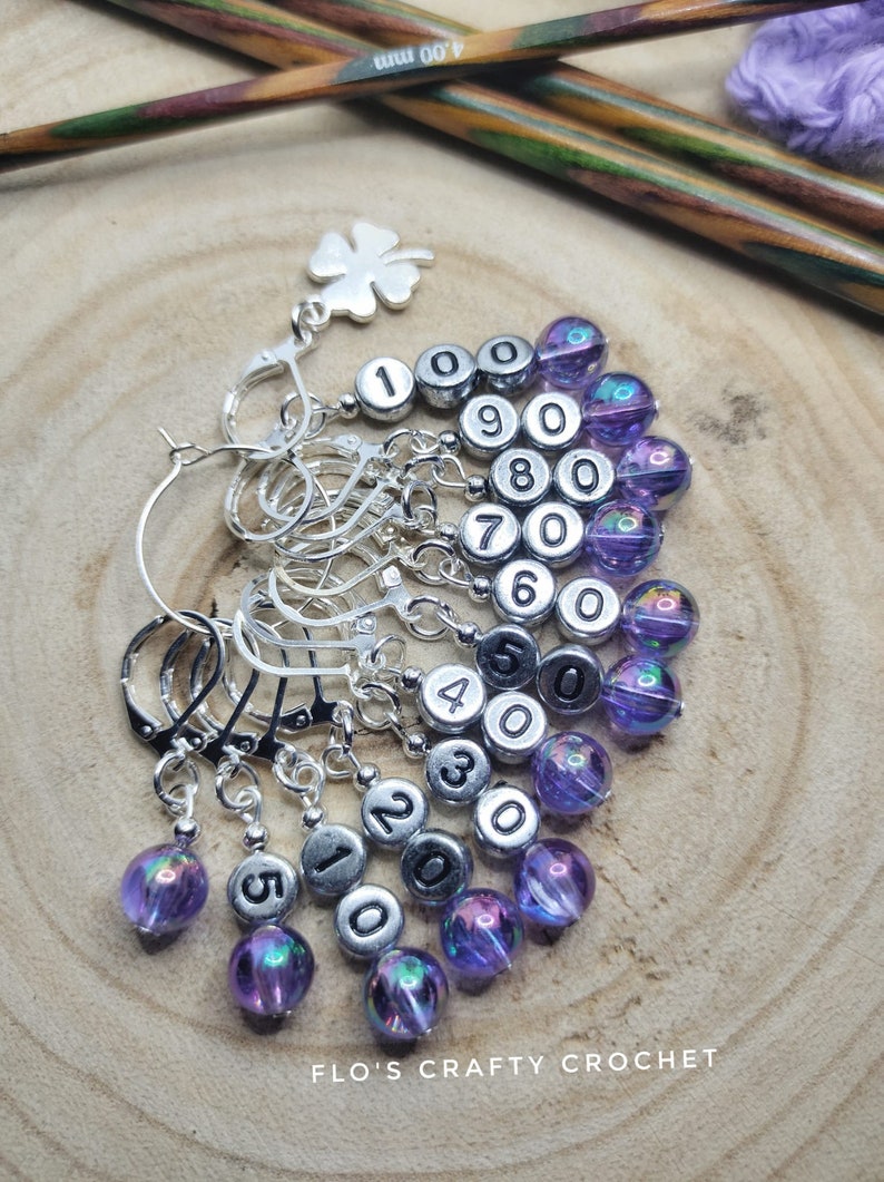 May include: A set of ten silver stitch markers with purple iridescent beads. Each marker has a silver ring and a bead with a black number from 1 to 10. The markers are arranged in a fan shape on a wooden surface.