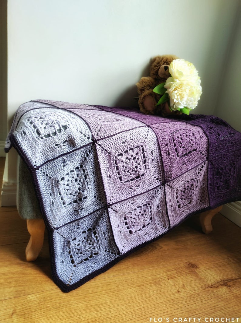 May include: A crocheted blanket with a gradient of purple and white squares. The blanket is draped over a wooden stool with a teddy bear holding a bouquet of white flowers.