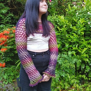Peut inclure: Un haut en crochet color&eacute; avec un motif ajour&eacute;. Le haut est port&eacute; sur un d&eacute;bardeur blanc et un jean noir. Le mod&egrave;le est debout devant un buisson vert avec des fleurs roses.
