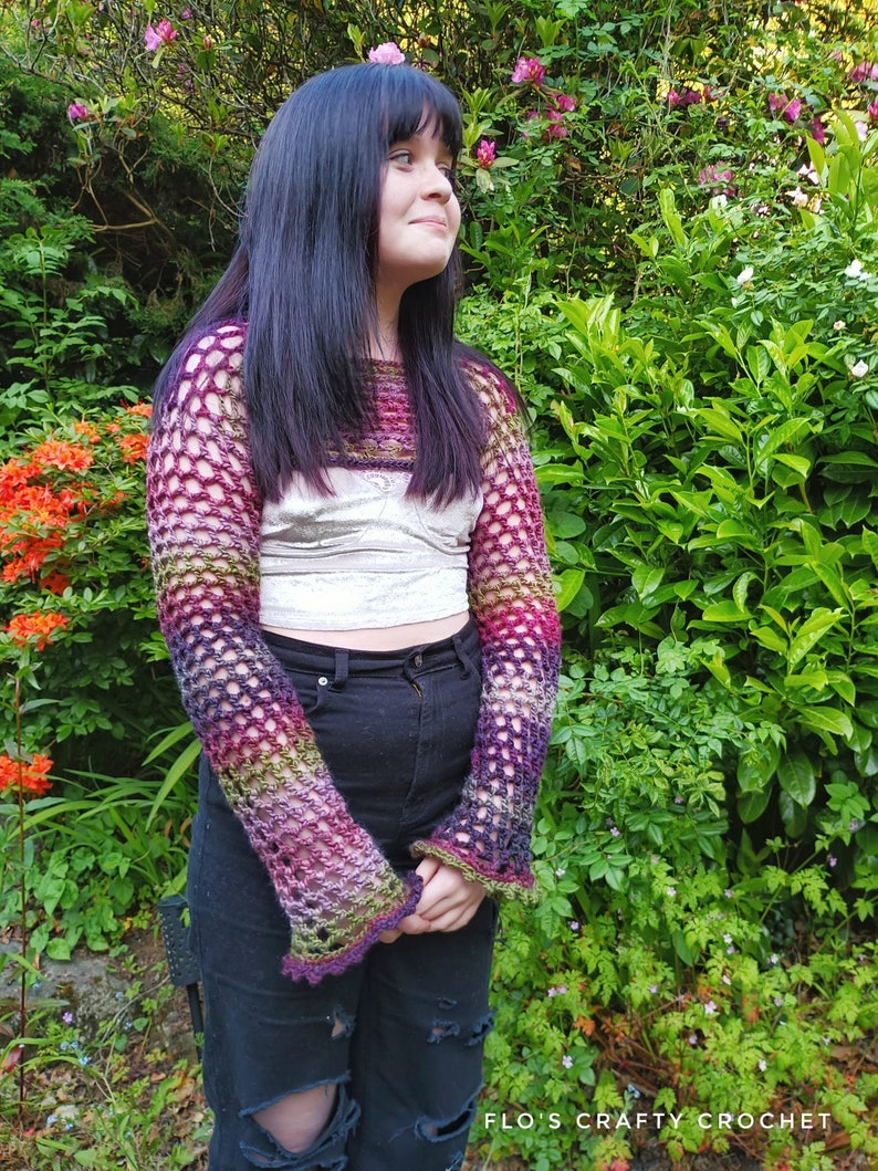 Peut inclure: Un haut en crochet color&eacute; avec un motif ajour&eacute;. Le haut est port&eacute; sur un d&eacute;bardeur blanc et un jean noir. Le mod&egrave;le est debout devant un buisson vert avec des fleurs roses.