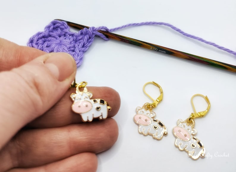 May include: A close-up of crochet supplies, including a purple crocheted flower, a wooden crochet hook, and a pair of gold-colored cow earrings with white enamel and black spots. The earrings have pink faces and are on a white background.