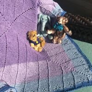 May include: A hand-knitted baby blanket in shades of purple and blue with a lacy border. The blanket is draped over a chair with three soft toys sitting on it.