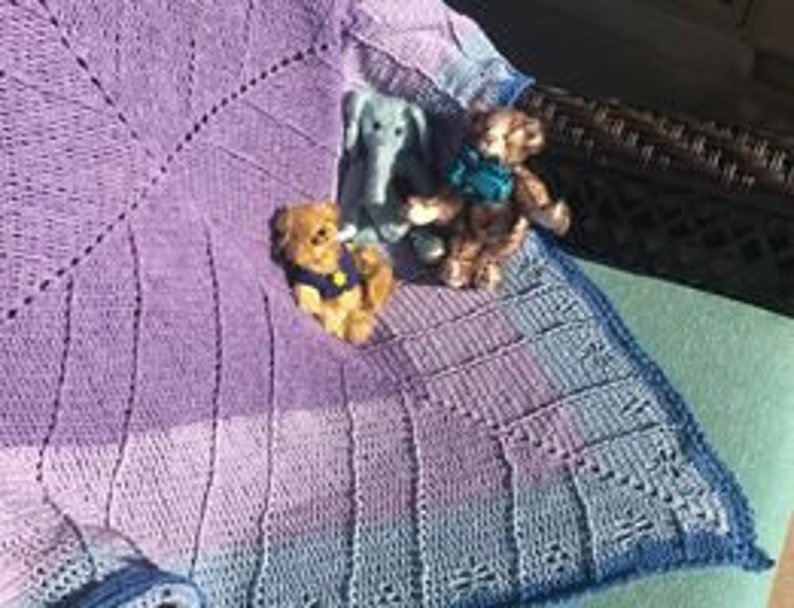May include: A hand-knitted baby blanket in shades of purple and blue with a lacy border. The blanket is draped over a chair with three soft toys sitting on it.