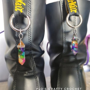 May include: Black leather boots with colorful crystal charms hanging from the top loops. The charms are multi-colored, with a silver metal cap and loop. The boot straps have the text "Dr. Martens" in yellow.
