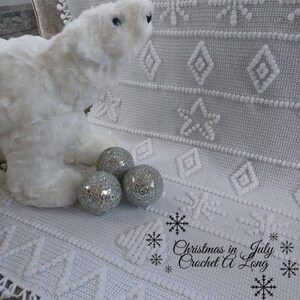 K&ouml;nnte beinhalten: Wei&szlig;e H&auml;keldecke mit Schneeflockenmuster und einem wei&szlig;en Pl&uuml;sch-Eisb&auml;ren. Im Vordergrund befinden sich drei silberne Dekorkugeln. Der Text "Christmas in July Crochet A Long" befindet sich in der unteren rechten Ecke.