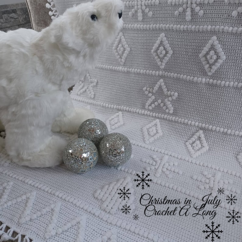 K&ouml;nnte beinhalten: Wei&szlig;e H&auml;keldecke mit Schneeflockenmuster und einem wei&szlig;en Pl&uuml;sch-Eisb&auml;ren. Im Vordergrund befinden sich drei silberne Dekorkugeln. Der Text "Christmas in July Crochet A Long" befindet sich in der unteren rechten Ecke.