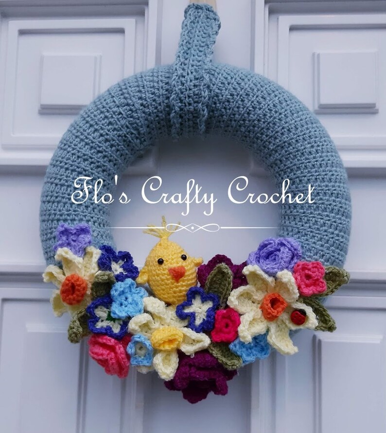 May include: A handmade crochet wreath in shades of blue, adorned with colorful crochet flowers and a yellow chick. The wreath features the text "Flo's Crafty Crochet". This decorative wreath is perfect for spring and Easter.