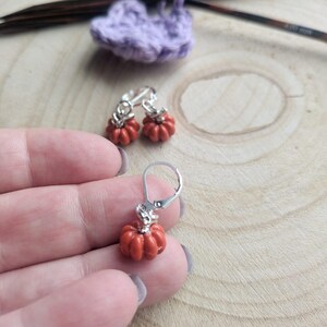May include: Small orange pumpkin earrings with silver leverback fastenings. The earrings have a detailed, textured surface. A crocheted purple heart and knitting needles are in the background. The text "FLO'S CRAFTY CROCHET" is visible.