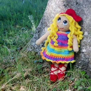 Könnte beinhalten: Eine gehäkelte Puppe mit langen blonden Haaren, die ein regenbogenfarbenes Kleid und rote Schuhe trägt. Die Puppe sitzt an einem Baumstamm.