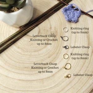 May include: Close-up of stitch markers with different fastenings, including leverback clasps and lobster clasps. The markers are available in silver or gold and are suitable for knitting or crocheting projects up to 8 mm in diameter. The image also shows knitting needles and a crocheted flower.