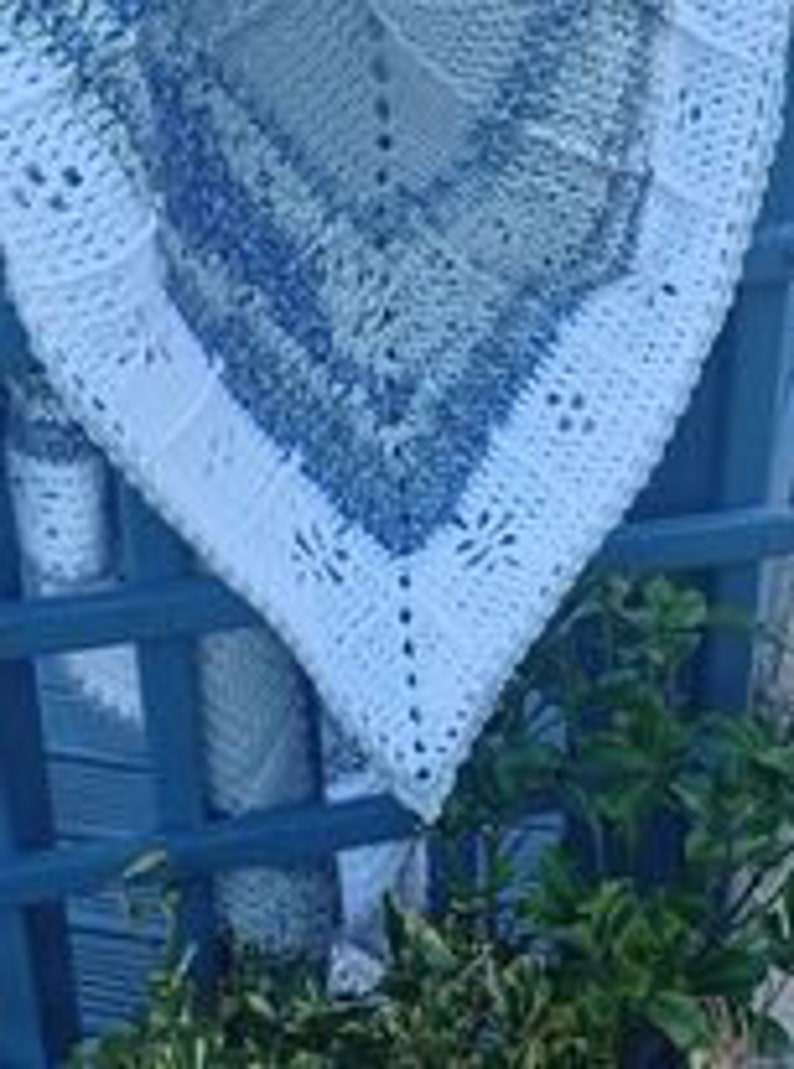 May include: A white crocheted baby blanket with a blue and white pattern. The blanket is folded and draped over a blue wooden fence.