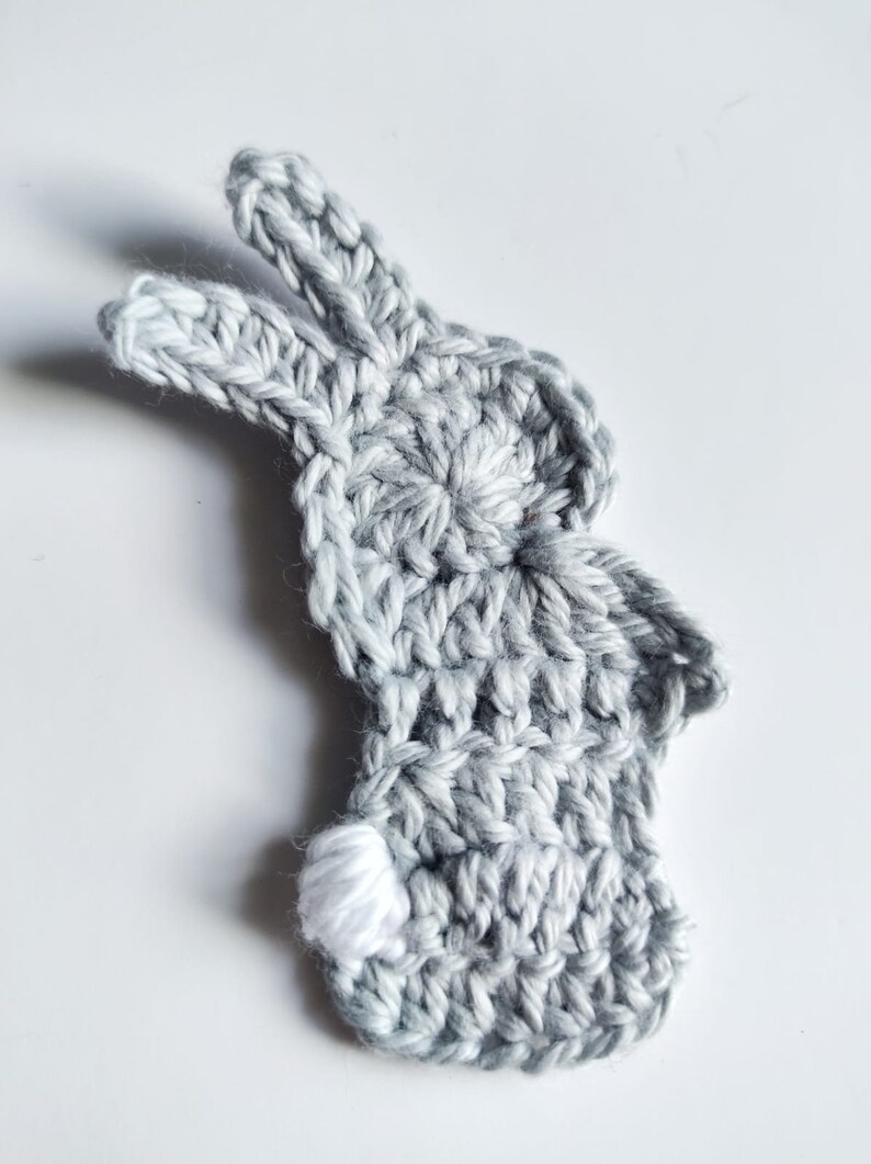 May include: A gray crocheted bunny with a white crocheted tail. The bunny is a flat, two-dimensional shape.
