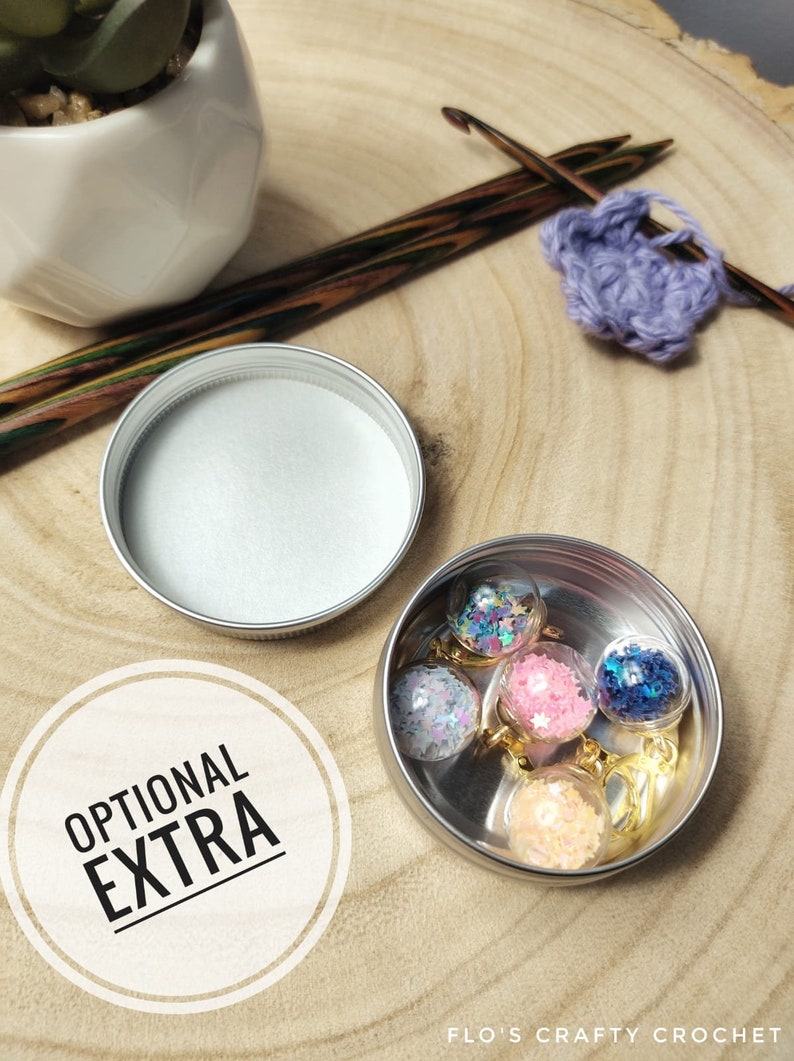 May include: A collection of small, clear glass ball charms filled with colorful glitter and star-shaped confetti, displayed in a silver tin. The charms are on gold-colored keychains. The image also includes a container with white powder, crochet hooks, and the text "OPTIONAL EXTRA."