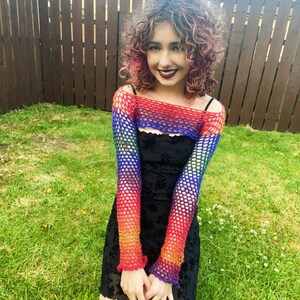 Peut inclure: Une personne portant un haut en crochet arc-en-ciel sur une robe noire. Le haut a des manches longues et un col large. La personne est debout dans une zone herbeuse.