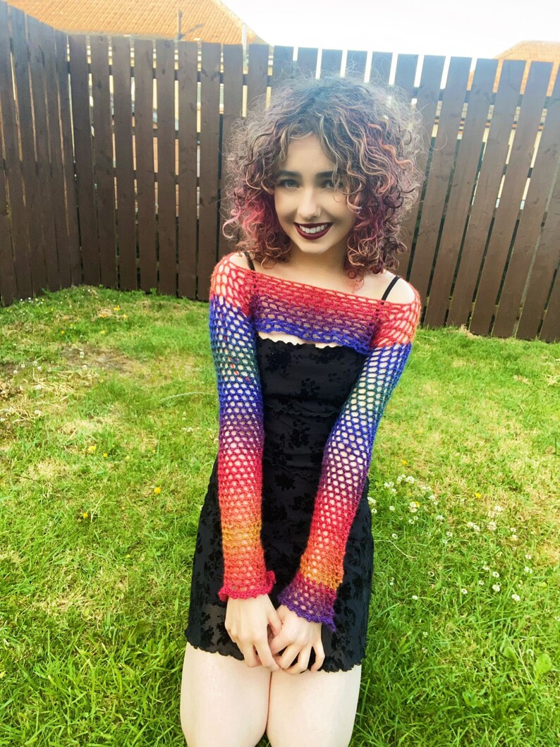Peut inclure: Une personne portant un haut en crochet arc-en-ciel sur une robe noire. Le haut a des manches longues et un col large. La personne est debout dans une zone herbeuse.