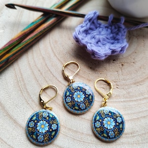 May include: Three gold stitch markers with blue and white floral designs. The markers are round and have a gold hook on the top.