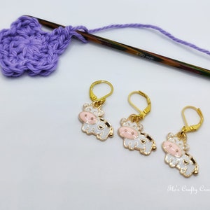 May include: Three gold-colored cow-shaped stitch markers with white enamel, black spots, and pink faces. A partially crocheted purple flower, a wooden crochet hook, and purple yarn are also in the image. The hook is marked "4.00 mm".