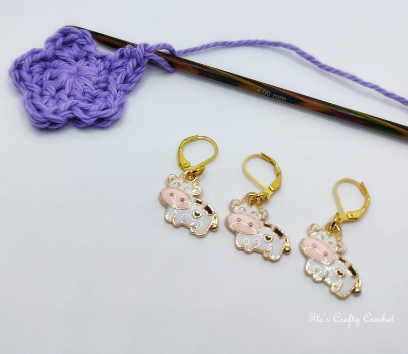 May include: Three gold-colored cow-shaped stitch markers with white enamel, black spots, and pink faces. A partially crocheted purple flower, a wooden crochet hook, and purple yarn are also in the image. The hook is marked "4.00 mm".