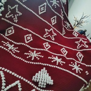 K&ouml;nnte beinhalten: Eine rote Wolldecke mit einem wei&szlig;en Schneeflocken- und Baummuster. Die Decke ist aus einem weichen, flauschigen Material gefertigt und hat eine strukturierte Oberfl&auml;che.