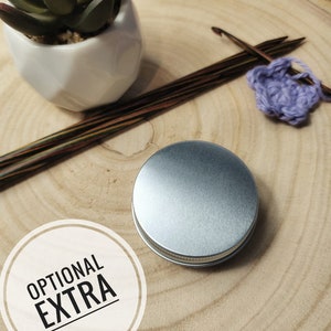 May include: A silver metal tin with the words "OPTIONAL EXTRA" printed on a white circle. The tin is sitting on a wooden surface with knitting needles and yarn.