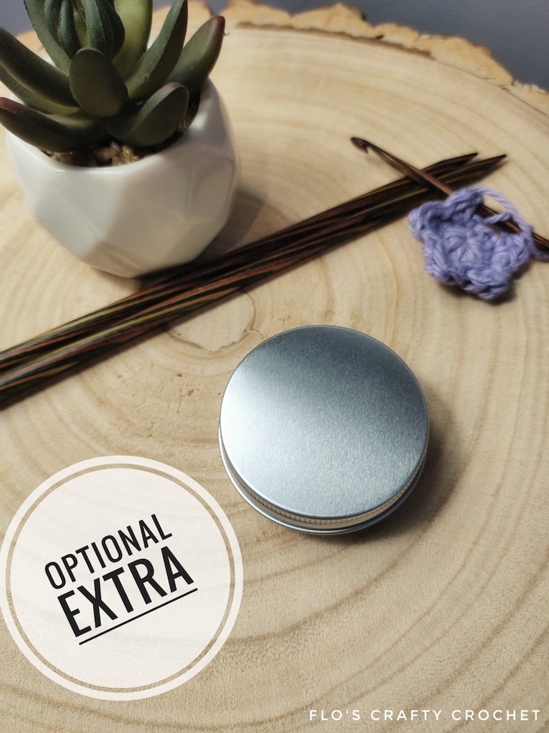 May include: A silver metal tin with the words "OPTIONAL EXTRA" printed on a white circle. The tin is sitting on a wooden surface with knitting needles and yarn.