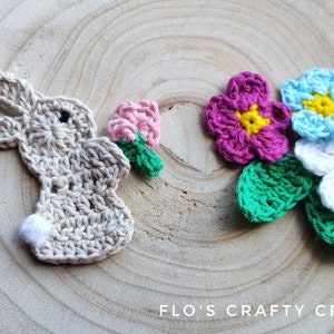 May include: Crocheted bunny and flowers. The bunny is beige with a white tail and black eyes. The flowers are pink, blue, and white with yellow centers. The flowers have green leaves.