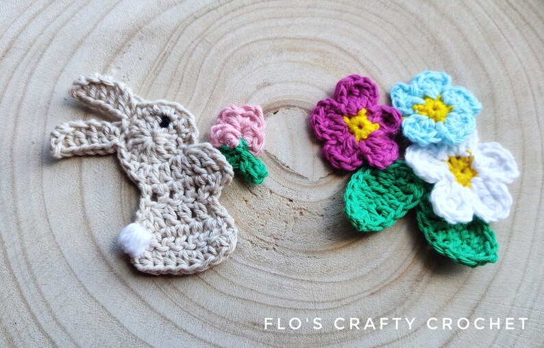 May include: Crocheted bunny and flowers. The bunny is beige with a white tail and black eyes. The flowers are pink, blue, and white with yellow centers. The flowers have green leaves.