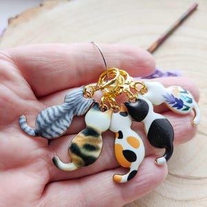 Pode incluir: Um conjunto de cinco marcadores de ponto em forma de gato em várias cores e padrões, incluindo cinza tigrado, tartaruga e preto e branco. Os marcadores estão presos a um anel dourado. O texto "FLO'S CRAFTY CROCHET" é visível.