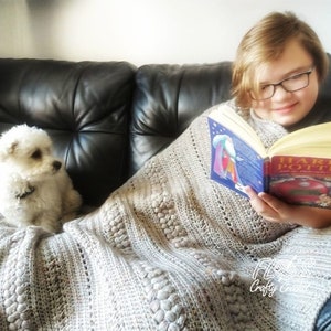 May include: A young person sits on a couch with a white dog and reads a book titled "Harry Potter and the Philosopher's Stone". They are both covered with a crocheted blanket.